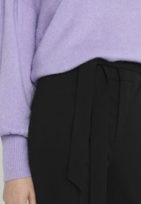 Levandulový pletený svetr s texturovaným povrchem, spárovaný s černými kalhotami s vysokým pasem, které mají zavazovací látkový pásek v pase.