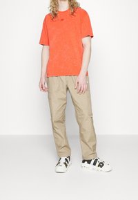 Oranžové tričko s tie-dye vzorem a malým logem, světlé béžové cargo kalhoty a bílé tenisky s černými a zlatými akcenty. Minimalistický design.