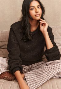 Frau, die im Schneidersitz auf einer beigen Couch sitzt, trägt einen dunkel gesprenkelten Pullover, braune Karohosen und braune Sneaker und berührt nachdenklich ihr Gesicht.