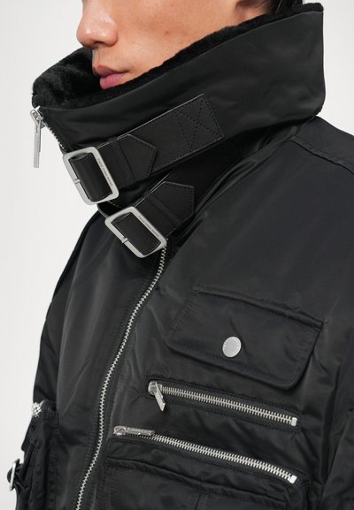 Chaqueta aislante negra con alto cuello, forro de piel, cremalleras plateadas y múltiples bolsillos. Presenta correas ajustables con hebillas de metal.