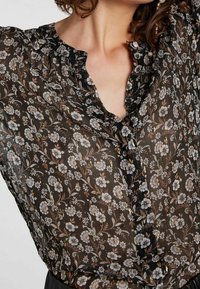 Zwarte, transparante blouse met een bloemenprint in wit en gouden accenten; heeft een ruchesnek, knoopsluiting en een subtiele textuur.