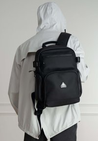 Mochila negra hecha de nylon duradero con un bolsillo frontal con cremallera, correas ajustables y un detalle del logo en forma de diamante.
