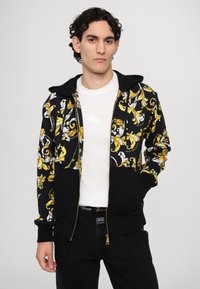 Κουκούλα ζακέτα με μαύρη βάση, περίπλοκα κίτρινα και λευκά λουλούδια, και μπροστινές τσέπες. Συνδυάζεται με λευκό μπλουζάκι από κάτω.