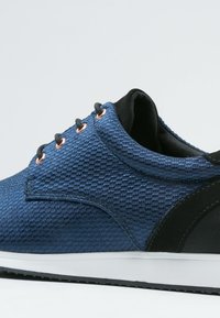 Gros plan d'une basket texturée bleue avec des lacets noirs, des œillets en cuivre, un soutien talon noir et une semelle blanche sur un fond gris clair.