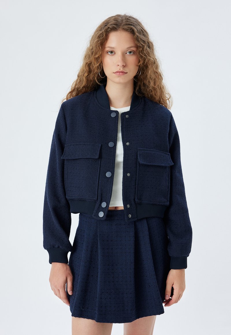 Marineblaue, kurze Bomberjacke mit zwei Vorderaschen, gerippten Bündchen und strukturiertem Stoff, kombiniert mit einem passenden Plisseerock.