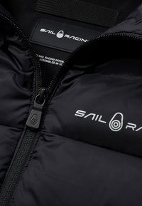 Sort pufferjakke med frontlynlås, der har en glat tekstur, "SAIL RACING" logo i hvidt og en stofetiket indeni.