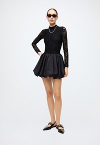 Black lace long-sleeve top with a high neckline, paired with a voluminous black satin skirt. Black flat shoes with straps complete the look.