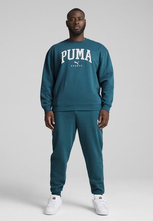 Türkiser Sweatshirt und passende Hose, beide mit weißem "PUMA"-Logo. Glatte Textur, entspannte Passform und weiße Sneakers runden das Outfit ab.