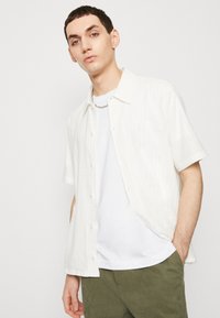 Kortärmad, knäppt skjorta i vit texturerad tyg, bärs över en enkel vit t-shirt. Modellen står mot en enkel bakgrund.