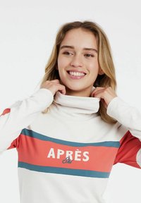 Langärmliges, cremefarbenes Oberteil mit hohem Kragen, das rote und blaue gestreifte Akzente sowie den Text "APRÈS Ski" auf der Vorderseite aufweist. Glatte Textur.