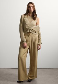 Top beige lavorato a maglia con spalle scoperte, caratterizzato da un dettaglio con bottoni; abbinato a pantaloni leggeri beige chiaro, a vita alta e larghi. Superfici texture lisce.