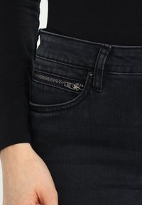 Tmavé džínové kalhoty s zipovou kapsou, hladkou texturou a úzkým střihem, přičemž částečně viditelná je černá košile s dlouhým rukávem.