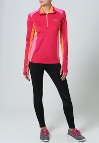 Różowo-pomarańczowa sportowa bluza z długim rękawem i suwakiem do ćwiartki, zestawiona z czarnymi legginsami i szarymi butami sportowymi z różowymi akcentami.