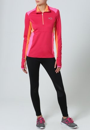Růžovo-oranžové sportovní triko s dlouhým rukávem a částečným zipem, doplněné černými legínami a šedými teniskami s růžovými prvky.