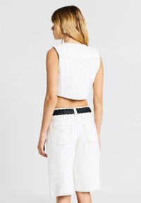 Donna con capelli biondi, che indossa un top bianco senza maniche, pantaloni bianchi fino al ginocchio e una cintura intrecciata nera, in piedi di spalle.