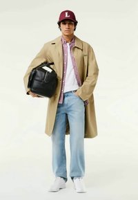 Jovem usando um casaco bege, camisa xadrez vermelha, jeans azul claro, tênis brancos, boné vermelho, segurando uma bolsa preta debaixo do braço.