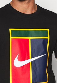 Camiseta negra de algodón con un gran estampado gráfico en rojo, azul y verde con un borde amarillo; incluye un logo blanco de Nike en la base.