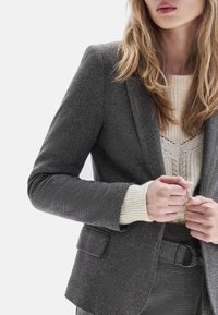 Mujer vestida con un blazer gris texturizado sobre un suéter de punto crema, ajustando el blazer con ambas manos visibles, cabello suelto y ondulado.