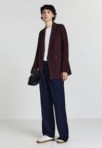 Donna che indossa un blazer bordeaux scuro, una camicia bianca, pantaloni larghi a righe navy, scarpe da ginnastica bianche, mentre tiene una pochette nera.