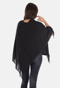 Poncho nero lavorato a maglia con texture a coste, caratterizzato da bordi frangiati e una vestibilità ampia, si adagia sulle spalle, presentato da dietro.