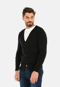Cardigan nero realizzato in materiale morbido, con design a V, chiusura a bottoni e polsini a coste. Abbinato a una maglietta bianca e jeans blu.