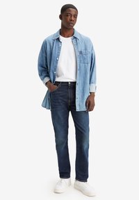 Camisa de mezclilla azul clara sobre una camiseta blanca, combinada con jeans azul oscuro y zapatillas blancas. Corte recto con un detalle de bolsillo en la camisa.