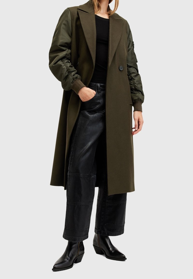 Abrigo verde oliva con mangas largas, diseño oversize, con un solo botón, combinado con pantalones de cuero negros de talle alto y botas puntiagudas.