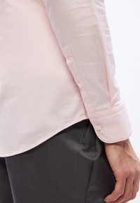 Το χέρι ενός άνδρα που ακουμπά κοντά στην τσέπη, φοράει ανοιχτό ροζ μακρυμάνικο πουκάμισο με μανσέτα με κουμπιά και σκούρο γκρι παντελόνι.