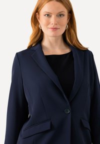 Blazer blu navy realizzato in tessuto elastico. Presenta una chiusura con un solo bottone, rever a punta e due tasche frontali. Texture liscia.