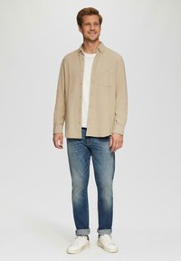 Beige Knopfenhemd, darunter ein weißes T-Shirt, blaue Jeans und weiße Sneaker. Das Hemd hat eine Brusttasche und lange Ärmel.