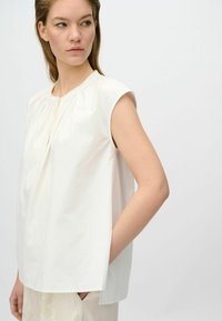 Blusa bianca a maniche corte con dettaglio plissettato sul davanti e scollatura rotonda. Tessuto morbido e vestibilità ampia. Tasche laterali visibili.