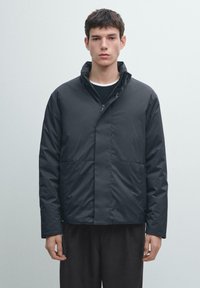 Schwarze gesteppte Jacke mit Stehkragen, ausgestattet mit einem Frontreißverschluss und zwei unteren Taschen. Glatte Textur und minimalistisches Design.