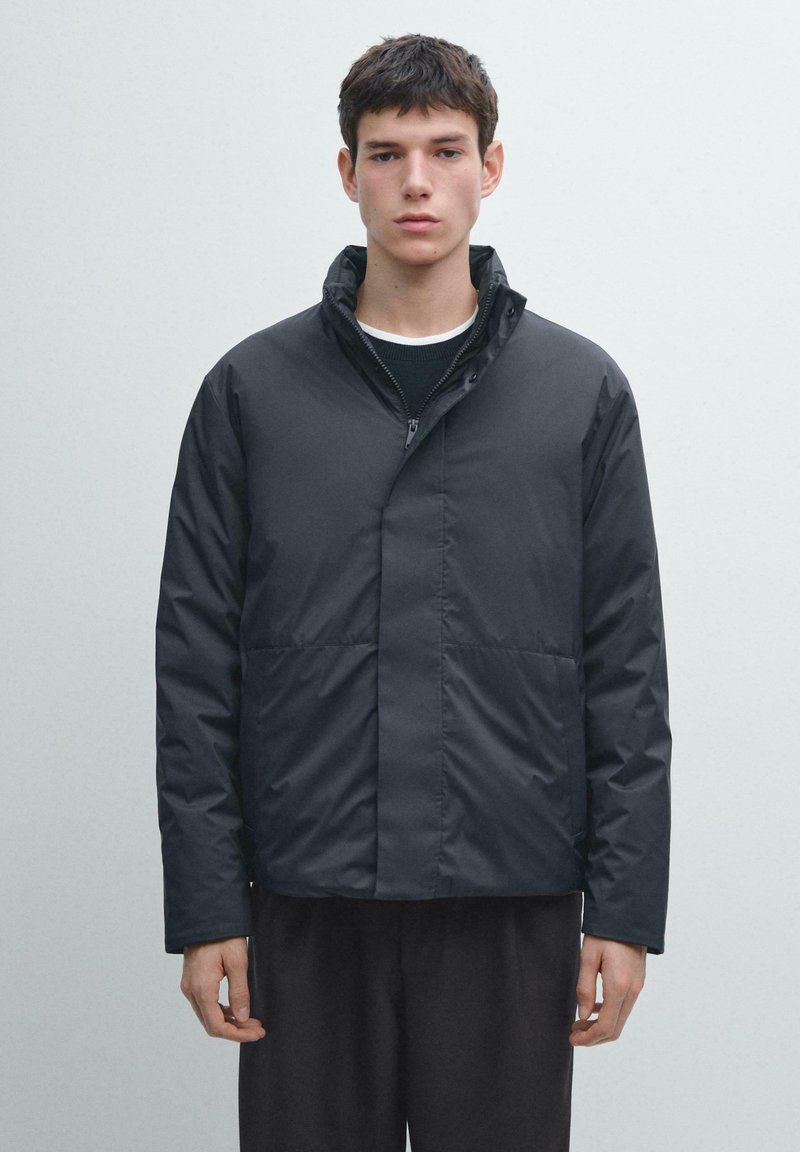 Schwarze gesteppte Jacke mit Stehkragen, ausgestattet mit einem Frontreißverschluss und zwei unteren Taschen. Glatte Textur und minimalistisches Design.