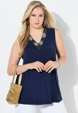 Blonde Frau in ärmellosem marineblauen Oberteil mit perlenbesetztem Ausschnitt, die eine goldene geflochtene Tasche hält und goldene Creolen sowie ein Armband trägt, vor weißem Hintergrund.