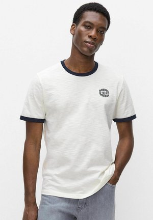 Jonge man met donkere huid, die een wit T-shirt met zwarte bies en kleine zwarte tekst op de borst draagt, staat met de hand in de zak van een lichtblauwe spijkerbroek.