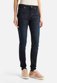 Jean skinny bleu foncé avec des coutures orange, des poches avant et une fermeture à boutons. Associés à des baskets rose clair avec des accents blancs.