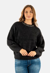 Suéter negro y esponjoso con un escote redondo adornado con acentos de cristal. Presenta mangas largas y un corte holgado, combinado con jeans azules.