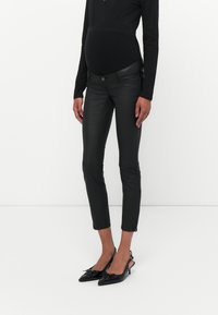 Pantalones ajustados de piel negra con cintura media, que cuentan con cremalleras laterales y costuras detalladas, combinados con tacones slingback negros.