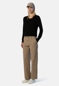 Schwarzer V-Ausschnitt-Pullover, beige gerade geschnittene Hose und weiße Sneakers. Begleitet von einer hellgrauen Mütze mit gesticktem Text.