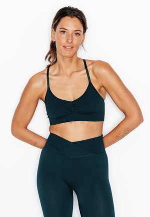 Donna con capelli castani raccolti indietro, che indossa un reggiseno sportivo verde petrolio scuro e leggings a vita alta, in piedi con le mani dietro la schiena.