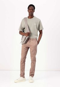 Camiseta de algodón verde claro, pantalones beige y zapatillas blancas. El modelo sostiene un bolso de hombro gris claro con un bolsillo con cremallera.