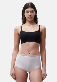 Frau mit langen braunen Haaren, die ein schwarzes Cropped-Top und weiße Unterwäsche mit hoher Taille und Spitzenbesatz trägt, steht vor einem schlichten Hintergrund.