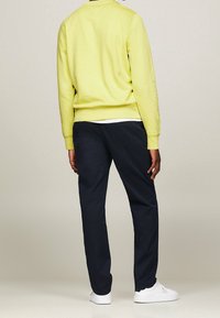 Žlutá mikina s kulatým výstřihem, v kombinaci s tmavě modrými kalhotami. Outfit je doplněn bílými teniskami. Materiál působí měkce a neformálně.