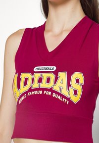 Bordový bezrukávový crop top s nápisem "ADIDAS" velkými žlutými písmeny a černým akcentem, vyrobený z měkkého, elastického materiálu.