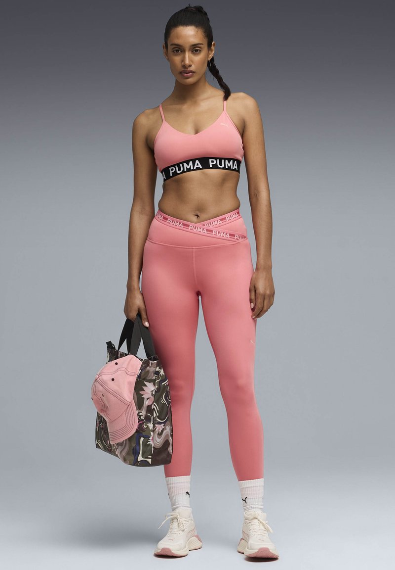 Γυναίκα με ροζ αθλητικό σουτιέν και κολάν Puma, κρατώντας μια τσάντα tote με σχέδια και ροζ καπέλο, φοράει λευκά αθλητικά παπούτσια Puma και κάλτσες.