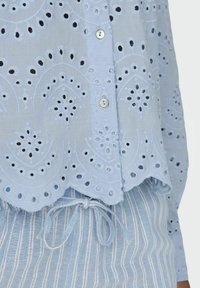 Camisa azul claro con bordados y cortes, presentando un dobladillo con forma de onda y detalles de botones. Combinada con una tela de rayas azul claro debajo.