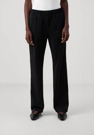 Personne portant un pantalon noir droit avec une taille élastique et des chaussures noires pointues sur un fond clair uni.