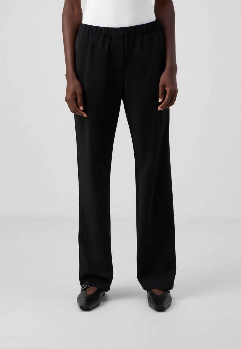 Personne portant un pantalon noir droit avec une taille élastique et des chaussures noires pointues sur un fond clair uni.