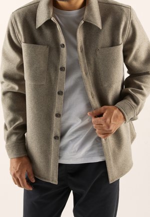 Mann trägt eine beigefarbene, geknöpfte Wolljacke über einem weißen T-Shirt und schwarzen Hosen und richtet die Jacke mit der rechten Hand.