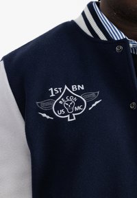Námornícky vlnený varsity kabát s bielymi rukávmi, obsahujúci predné logo s "1st BN", dizajnom srdca a detailom lebky. Pruhovaný golier.